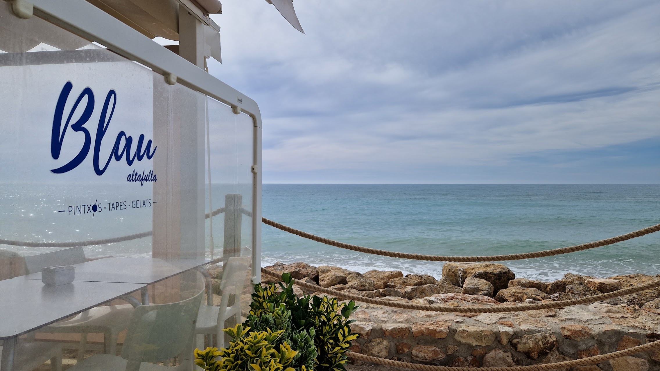 Vista del océano desde una terraza, con una cuerda de protección y el cartel "Blau altafulla" en una mampara transparente.